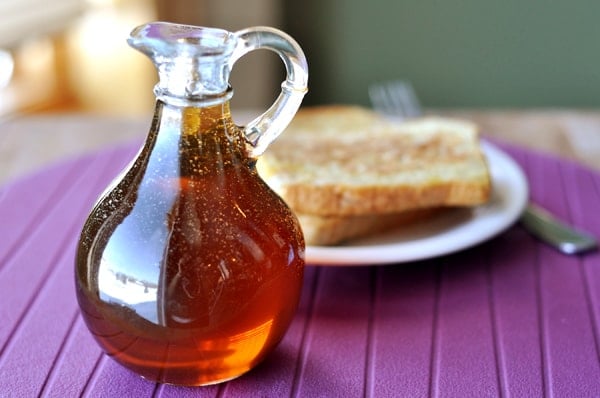 A jug of maple syrup in front of a plate of french toast.