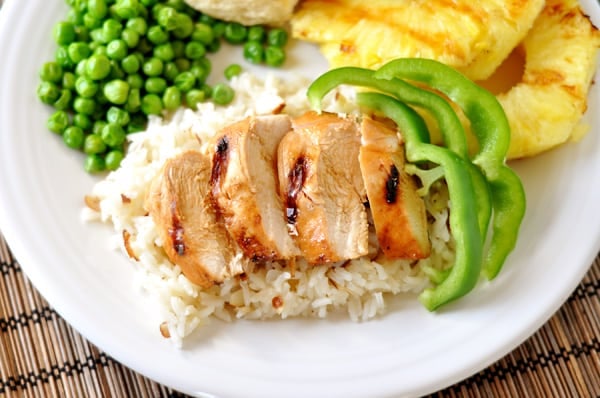 Top down view of a white plate full of white rice with chicken slices on top, green pepper slices to the side, green peas, and grilled pineapple slices.