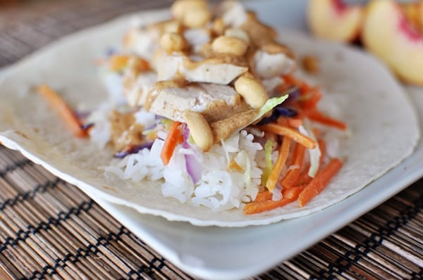 White tortilla topped with cooked rice, asian chicken, veggies, and peanuts stacked on top.