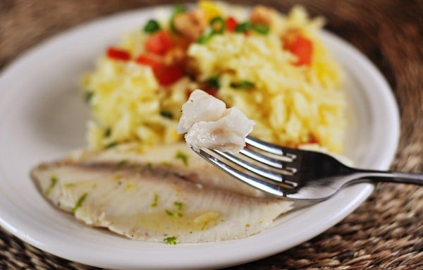 White plate with cooked tilapia and one bite of tilapia on a fork, and a serving of orange jasmine rice behind it.