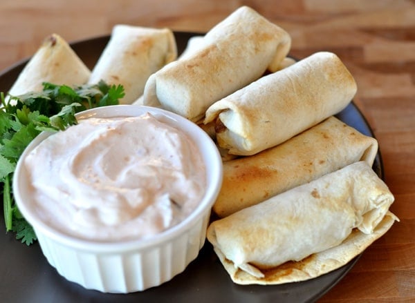 Golden brown cooked egg rolls surrounding a ramekin of dipping sauce on a black plate.