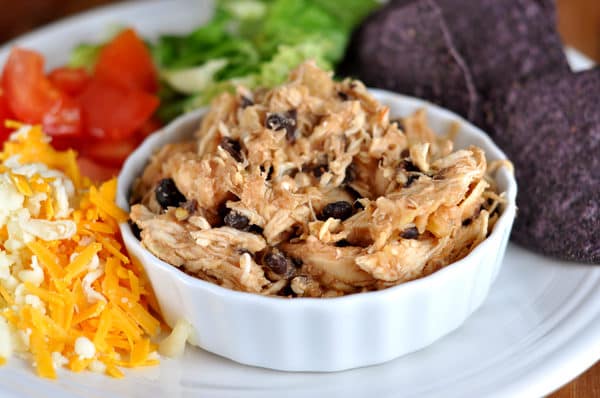 Small white ramekin with shredded chicken and black beans on a plate surrounded with toppings.