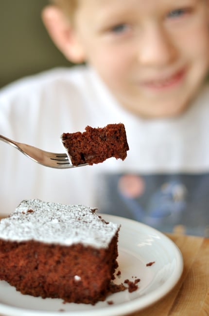 A piece of powdered sugar dusted cake with a little boy behind it holding a bite of the cake on his fork.