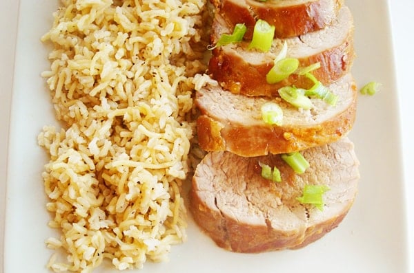 Sliced pork tenderloin next to brown rice on a white platter.