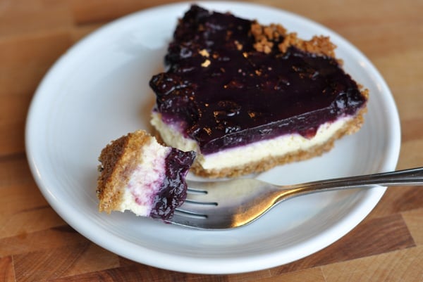 Slice of cheesecake with blueberry topping and a bite being taken out with a fork.