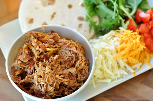 White platter with a bowl of shredded pork, flour tortillas, lettuce, tomatoes, and shredded cheese on it.