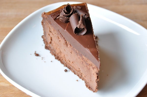 A thick slice of chocolate cheesecake with chocolate curls on top on a white plate.