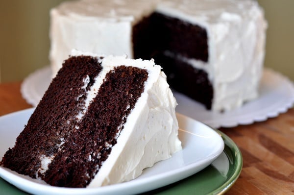 A big slice of two-tiered chocolate cake with white frosting on a white plate, with the rest of the cake behind it.