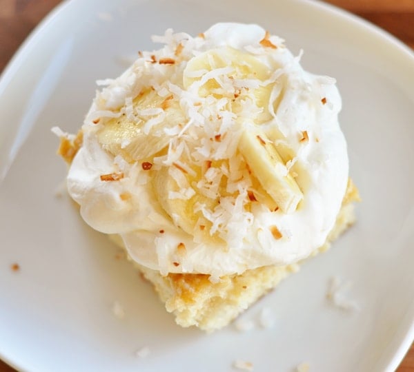 Top view of a white plate with a slice of tres leches cake topped with whipped cream and sliced bananas.