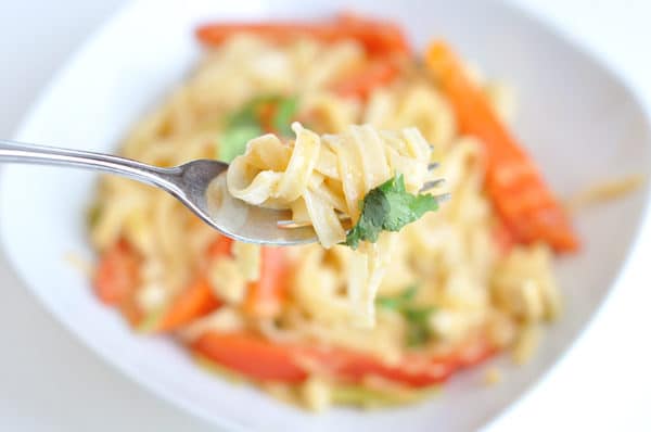 Top view of white bowl with cooked noodles and red pepper strips, with a fork above the bowl with a bite ready to be eaten.