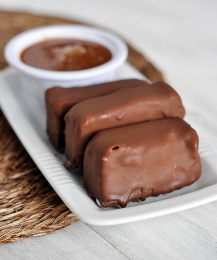 Three chocolate covered ice cream bars lined up next to each other on a white platter next to a bowl of caramel sauce.