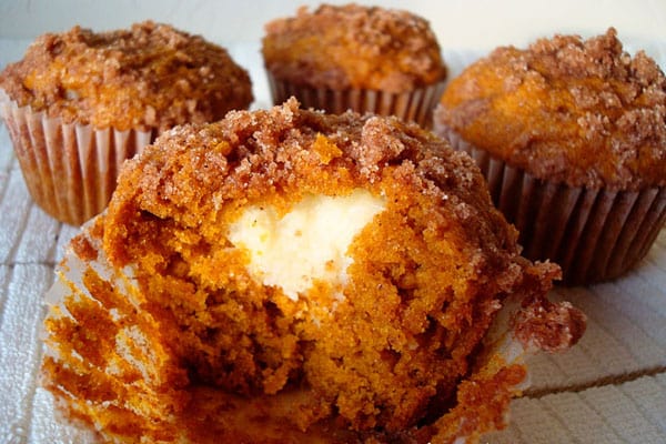 A cream cheese filled pumpkin muffin partially eaten in front of three other muffins.
