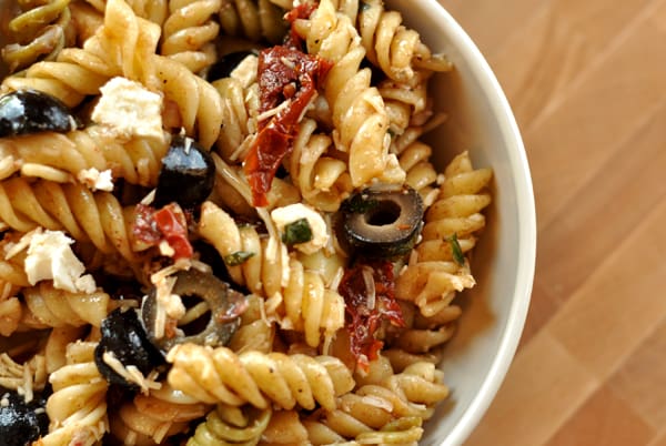 White bowl filled with cooked rotini, sliced olives, and sun-dried tomatoes.