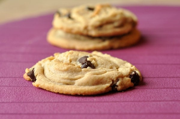 A chocolate chip cookie on a purple table, with two more cookies in the background.