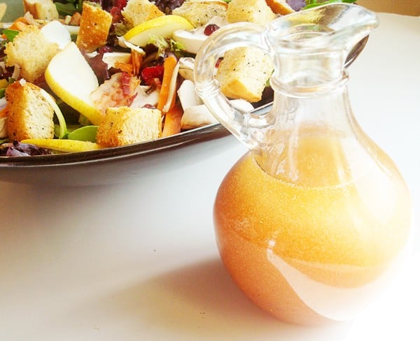 Small glass pitcher of salad dressing next to a bowl of salad.