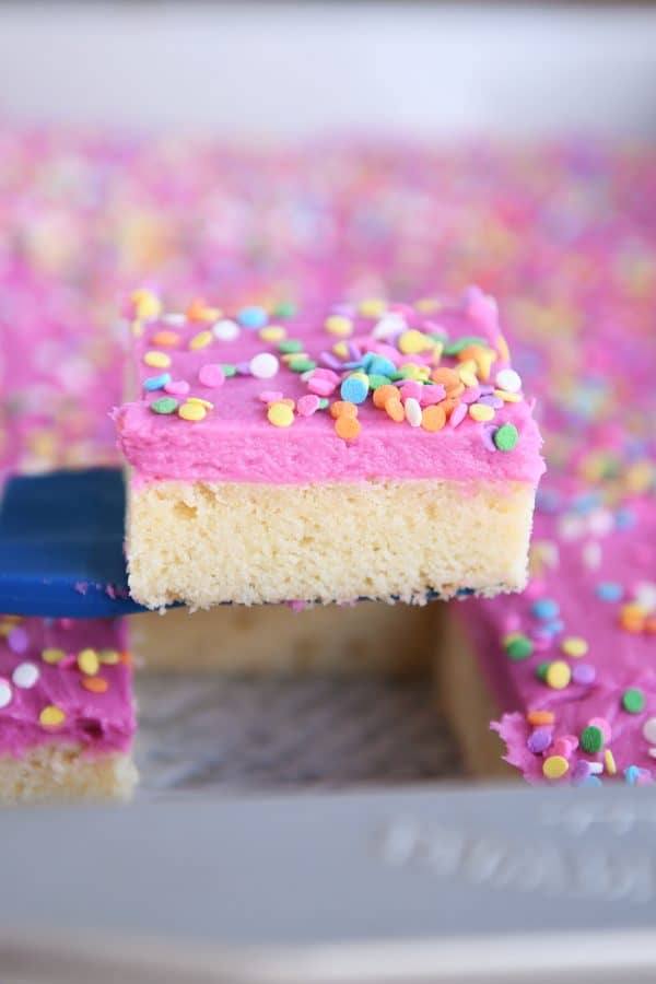 A blue spatula lifting a pink frosting and sprinkle-covered sugar cookie bar out of a metal pan.