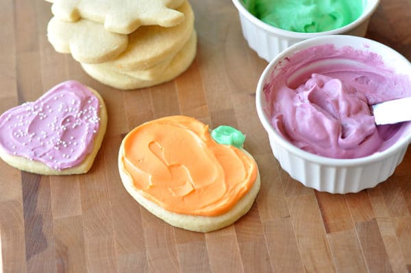 frosted heart and pumpkin sugar cookies, with ramekins of pink and green frosting and a stack of unfrosted sugar cookies all on a brown table