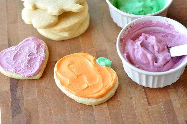 Frosted heart and pumpkin sugar cookies, with ramekins of pink and green frosting and a stack of unfrosted sugar cookies all on a brown table.