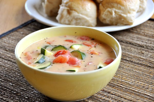 Bowl of soup filled with zucchini and tomatoes with a plate of rolls behind it.