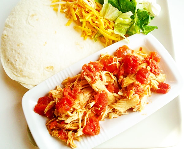 Shredded chicken and tomatoes in a white platter with white tortillas on the side.