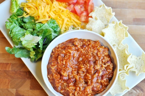 Top view of a white bowl with taco salad mixture and lettuce, cheese, tomatoes, and chips around the side.