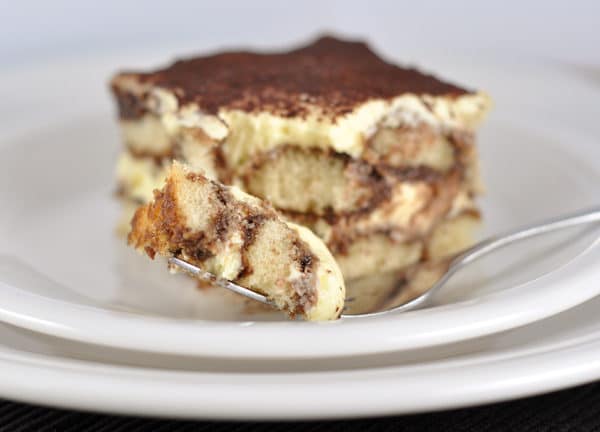 Piece of tiramisu on a white plate with a bite taken out of it.