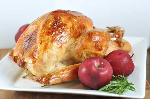 golden brown roasted whole turkey on a white platter with two red apples sitting beside it
