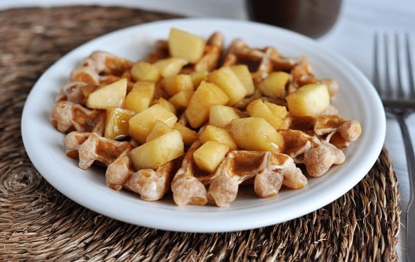 White plate with a waffle covered in apple chunks and a cinnamon sauce.