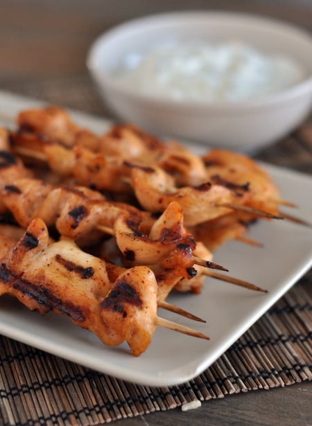 White platter with grilled chicken skewers with a bowl of dipping sauce behind it.