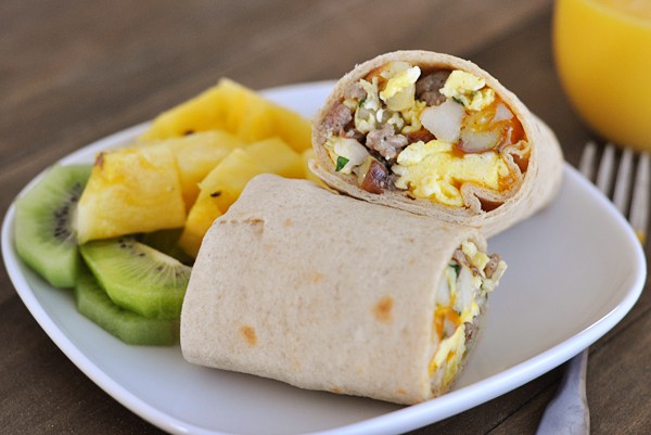 A breakfast burrito split in half on a white plate with fresh pineapple and kiwi on the side.