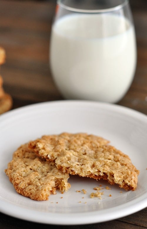A thin, baked oatmeal cookie split in half on a white plate, with a glass of milk in the background.