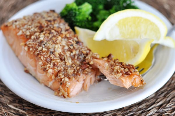 a white plate with a pecan crusted fillet of salmon, lemon wedges, and broccoli