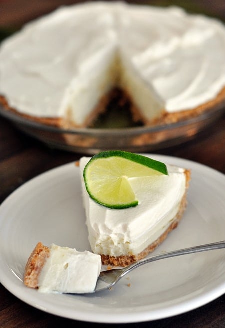 a slice of sour cream lime tart with a graham cracker crust on a white plate with the rest of the pie behind it