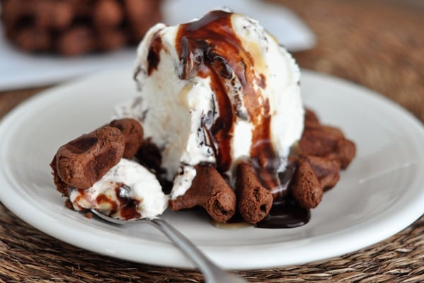 A chocolate waffle cookie topped with a scoop of vanilla ice cream and chocolate sauce on a white plate.