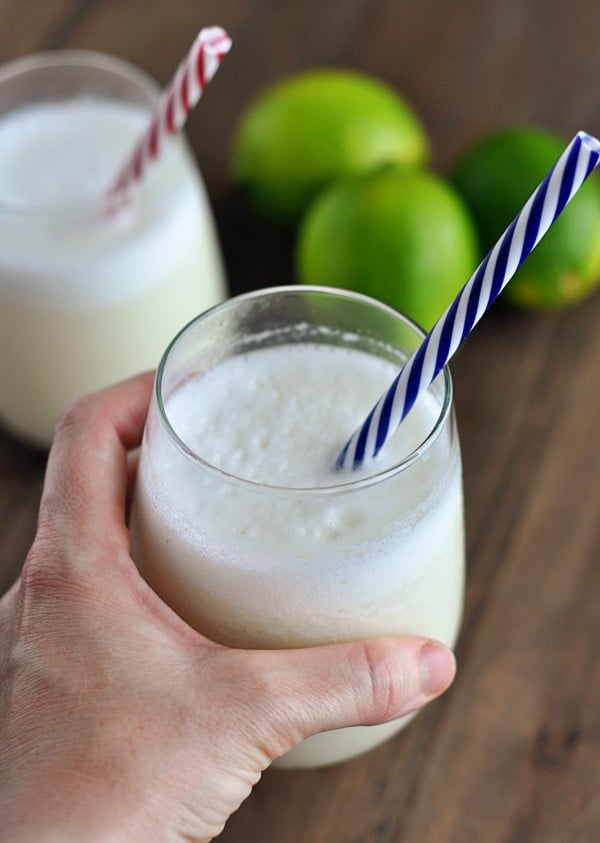 A glass of frothy lemonade with a blue and white striped straw in it and limes in the background.