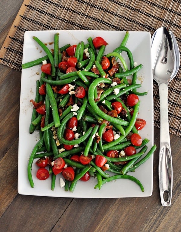 A top view of a white platter full of green beans, sliced cherry tomatoes, feta, and bacon sprinkled throughout. 