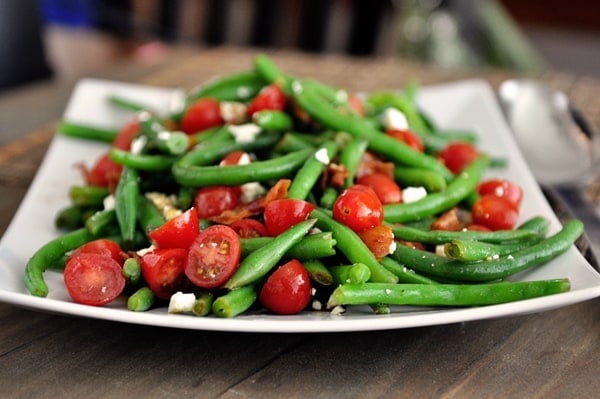 A white platter with cooked green beans, sliced cherry tomatoes, and feta cheese.