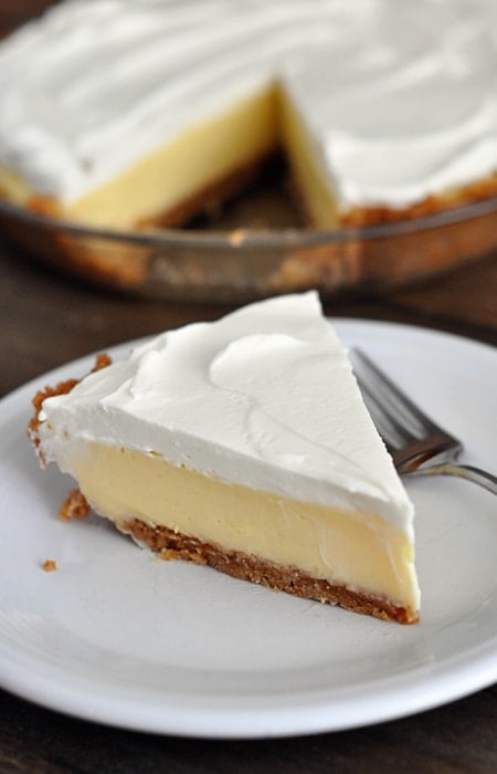 A slice of whipped cream topped, graham cracker crust lemon pie on a white plate with the rest of the pie in the background.