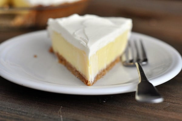 A slice of whipped cream topped, graham cracker crust lemon pie on a white plate.