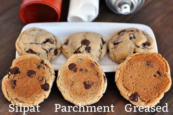 three bottom-up chocolate chip cookies lined up with the words silpat, parchment, and greased underneath and more cookies behind them