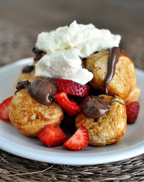 plate full of golden brown ebelskivers topped with sliced strawberries, chocolate sauce, and whipped cream