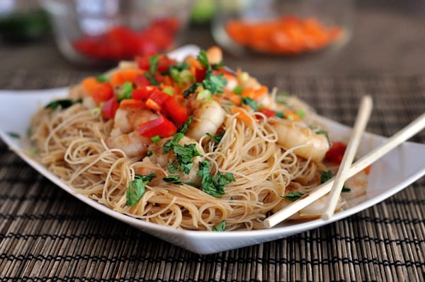 A white platter with cooked noodles, shrimp, vegetables and chopsticks on the side.