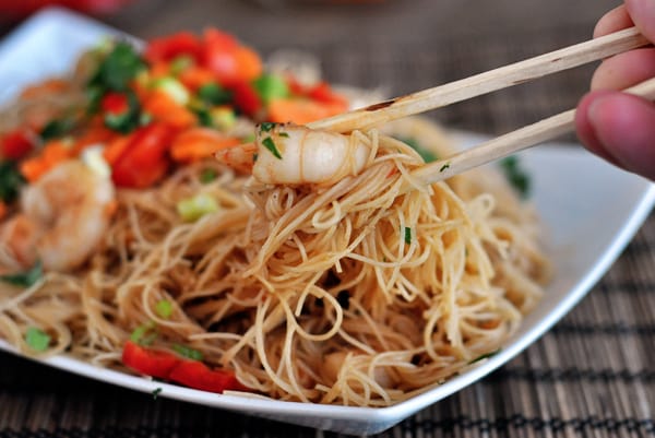 Chopsticks pinching a bite of noodles and shrimp from a white platter full of food.