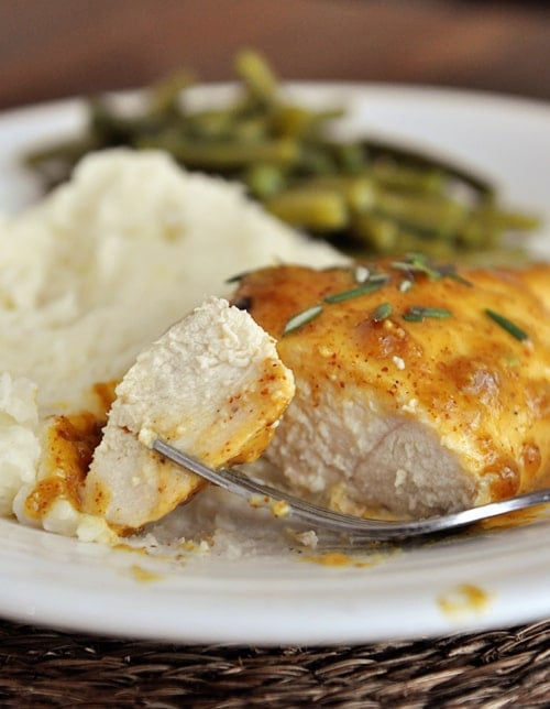 Mustard covered chicken breast with a bite cut next to mashed potatoes and green beans on a white plate.