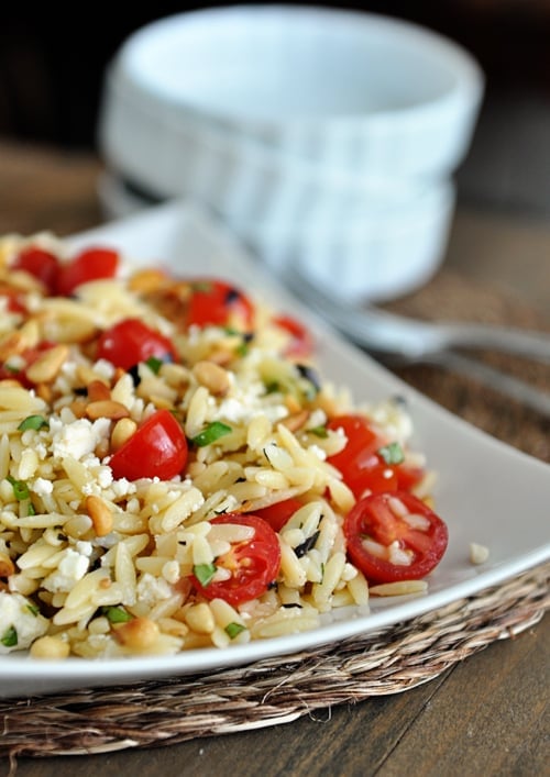 A white dish full of cooked orzo pasta, cherry tomatoes, pine nuts, and basil, and some white bowls and forks on the side.