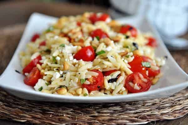 A front view of a white dish full of cooked orzo pasta, cherry tomatoes, pine nuts, and basil.