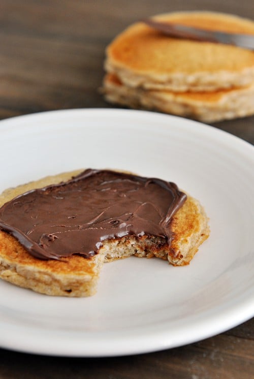 A pancake topped with Nutella, with a bite taken out, on a white plate.