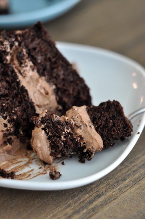 A piece of two-layer chocolate cake with chocolate frosting in the middle on a white plate, with one bite being taken out. 