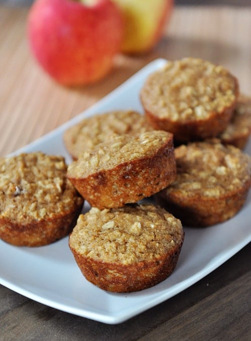 A white platter full of baked muffins, studded with oats, and apples in the background.