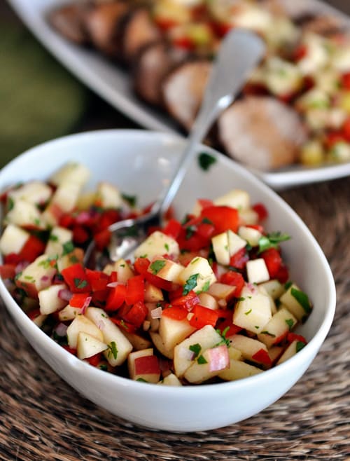 A white oval bowl with fresh apple salsa and sliced pork loin in the background.